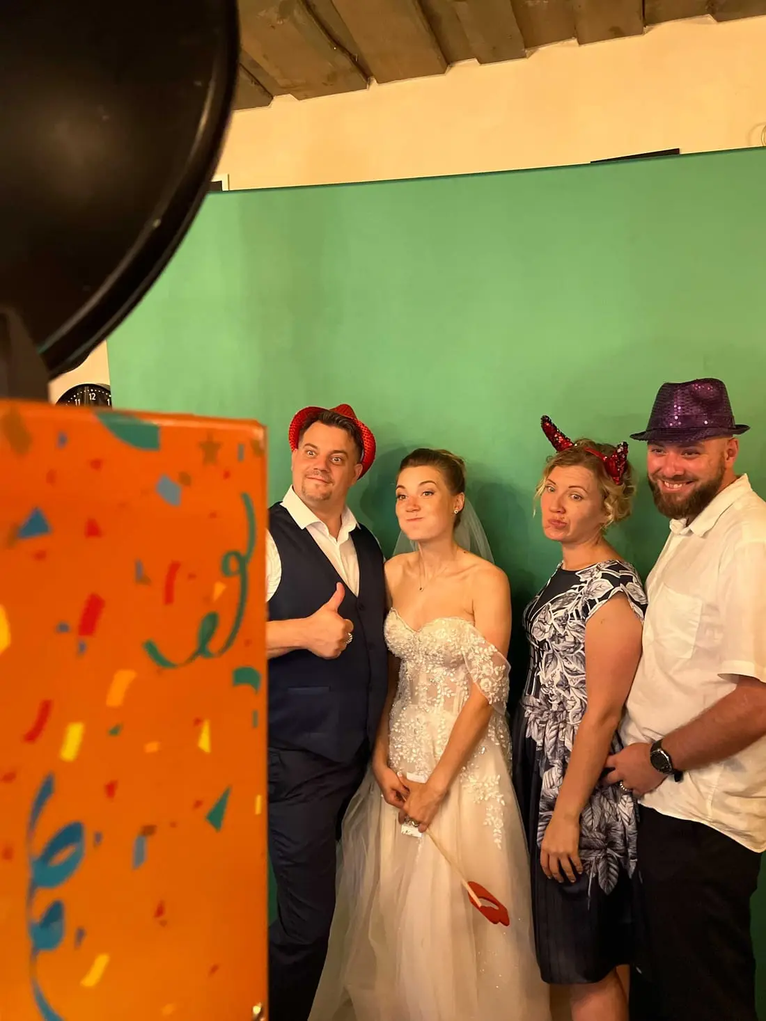 Līgava, līgavainis un viesi ar spīdīgām cepurēm un velna ragiem pozē foto kastei pie zaļa fona