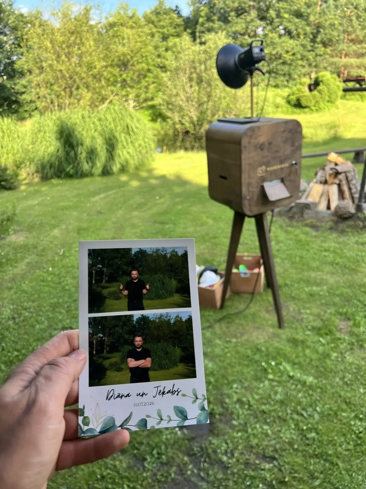 Rokā kāzu izdruka ar jaunā pāra vārdiem, fonā koka foto kaste zaļā dārzā