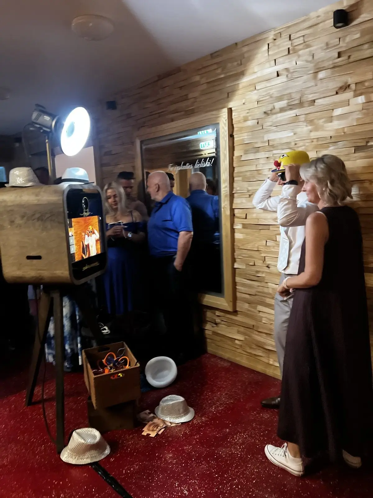 Koka foto kaste ar gredzenveida gaismu svinību telpā, uz grīdas rekvizītu kaste un cepures, viesi pozē pie koka sienas
